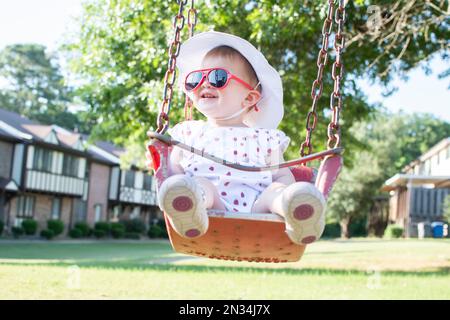 Süßes weißliches kleines Mädchen auf einer Schaukel mit roter Sonnenbrille Stockfoto