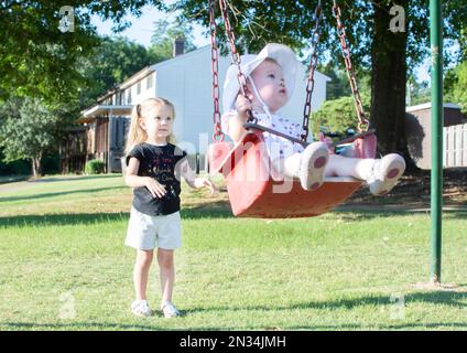 Die große Schwester hilft der kleinen Schwester. Geschwisterliebe. Kinder auf einer Schaukel Stockfoto