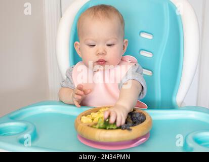 Baby isst Brokkoli. Säuglingsprothetik. Entwöhnung. Gesunde Ernährung. Kaukasisches Mädchen, das in einem Hochstuhl sitzt und ihr Mittagessen isst Stockfoto