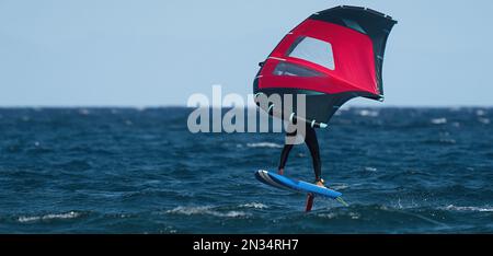 Ein Mann trägt Flügelfolien mit handgeführten aufblasbaren Flügeln und Tragflächensurfbrettern in einem blauen Ozean, reitet auf einem Windflügelbrett, surft auf den Wellen Stockfoto