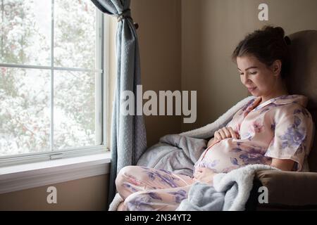 Eine schwangere weiße junge Frau, die im Schaukelstuhl neben dem Fenster sitzt, lächelt und ihren Bauch ansieht Stockfoto