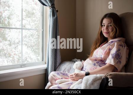Eine schwangere weiße junge Frau, die im Schaukelstuhl neben dem Fenster sitzt, lächelt und ihren Bauch ansieht Stockfoto