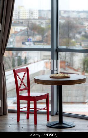 Pizza auf einem Holzbrett steht auf einem Tisch neben einem Fenster in einem Café. Hohe Panoramafenster im Restaurant. Ein Stuhl und ein Tisch neben einem großen Panorama Stockfoto