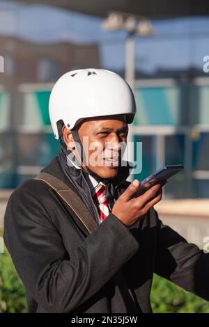 Ein dunkelhäutiger Geschäftsmann in einem Rollerhelm hält das Telefon und lächelt, während er über Voicemail spricht Stockfoto