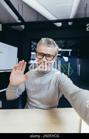 Ein erwachsener Mann im Büro, der mit der App am Telefon telefoniert, ein Geschäftsmann, der Spaß hat, mit Freunden und Kollegen zu sprechen, Selfie-Fotos zu machen. Stockfoto