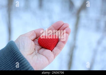 Rot gestricktes Herz in weiblicher Hand gegen den Schneewald. Konzept einer romantischen Liebe, Weihnachtsferien, Valentinstag, Winterwetter oder Wohltätigkeitsorganisation Stockfoto