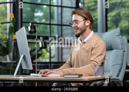 Ein junger männlicher Programmierer, Freiberufler, sitzt im Büro am Tisch neben dem Fenster und arbeitet am Computer und tippt auf der Tastatur. Lächelnd, mit Brille und beigefarbenem Pullover. Stockfoto