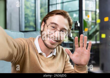 Nahaufnahme. Junger männlicher Blogger, Student, Geschäftsmann, der winkt, grüßt und mit der Kamera spricht. Am Schreibtisch im Büro sitzen, lächeln und Fotos machen. Stockfoto