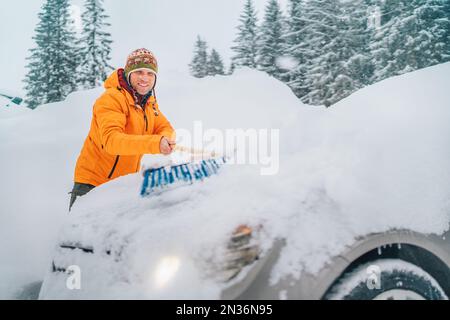 Lächelnder Mann in lustigem nepalesischen Hut, der Schnee von der Motorhaube entfernt, mit einer großen Schneebürste. Das Auto friert auf der Landstraße des Waldhauses Stockfoto