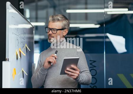 Ein erfahrener Geschäftsmann in der Nähe eines Whiteboards mit Diagrammen, die einen Strategieplan für die Finanzen des Unternehmens analysieren und darüber nachdenken, hält der Chef einen Tablet-Computer und einen Marker für Notizen in der Hand. Stockfoto