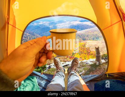 Heißes Getränk in der Hand und wunderbarer Blick auf die farbenfrohen Herbstberge durch den offenen Eingang zum Zelt. Die Schönheit einer romantischen Wanderung und Kamera Stockfoto