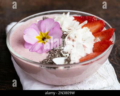 Erdbeerbananen Soja Jogurt Smoothie Schale, garniert mit Erdbeerscheiben, Chiasamen, Kokosflocken und essbarer primelblume Stockfoto