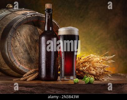 Ein Glas schwarzes Craft-Bier, eine Flasche Bier und ein Holzfass auf dem Holztisch im Keller. Stockfoto