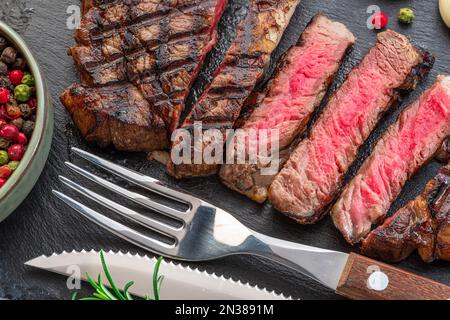 Gegrillte, köstliche Ribeye-Steak-Scheiben und Küchenutensilien auf schwarzem Schiefer-Servierteller. Flach verlegt. Stockfoto