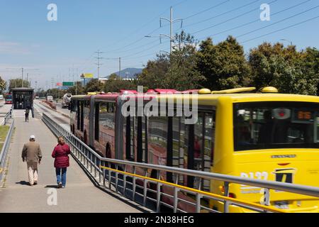 BOGOTA, KOLUMBIEN - zwei Personen gehen in eine Transmilenio-Station mit einem großen roten und gelben Bus in der Nähe Stockfoto