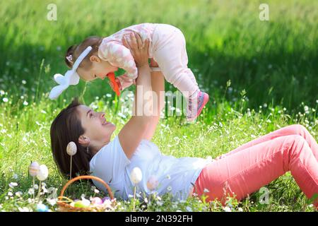 Mama zieht ihre kleine Tochter auf dem Rasen auf, um Spaß zu haben. Ein einjähriges Mädchen, das mit einer Frau spielt. Kleines Kind, mom feiert Ostern draußen in der Kammer Stockfoto