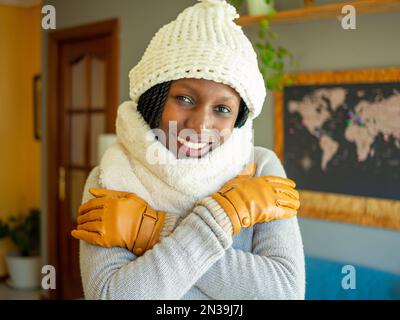 Eine junge Frau mit Handschuhen, Schal und Wintermütze, die in einem gemütlichen Wohnzimmer gegen die kalte Welle kämpft. Ich schaue in die Kamera Stockfoto