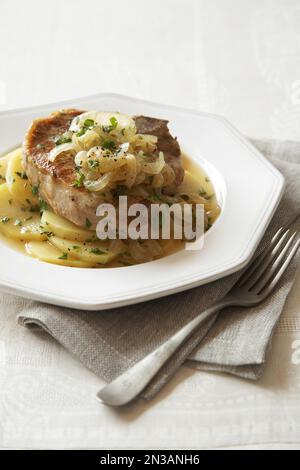 Schweinekotelett auf Kartoffelscheiben garniert mit Zwiebeln in einem achteckigen Gericht mit Gabel Stockfoto