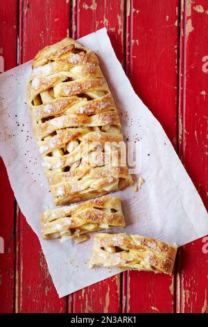 Apfelstrudel auf Pergamentpapier mit zwei abgeschnittenen Scheiben auf einer roten Stangenpappe Stockfoto