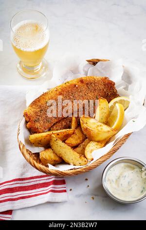 Panierte Tilapia mit Kartoffelkeilen in einem Korb und Tartesauce und ein Glas Bier Stockfoto