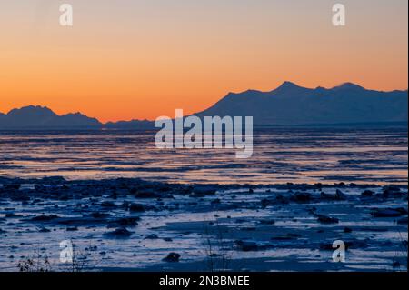 Wintersonnenuntergang aus der Innenstadt von Anchorage, Blick über den Einlass am Mt. Spurr und die Tordrillo Mountains in Silhouette, mit Eisbrocken und... Stockfoto