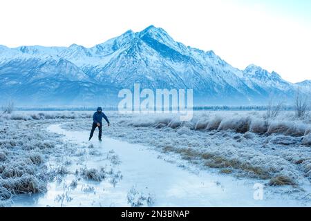 Kaukasischer Mann Hinterland Schlittschuhlaufen, nordic Blading, in der Wintersonne auf einem kleinen Bach gesäumt von frostbedecktem Gras mit Pioneer Peak und dem Chug... Stockfoto