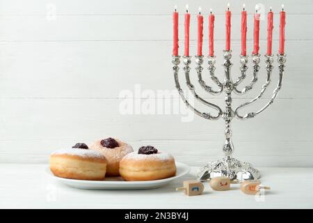 Silberne Menora, Dreidel mit He-, PE-, Nun-, Gimel- und Sufganiyot-Buchstaben auf weißem Holztisch, Platz für Text. Hanukka-Symbole Stockfoto