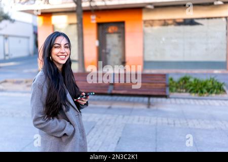 Positive junge Frau in warmen Kleidern, die am Tag mit ihrem Smartphone auf der Straße der Stadt steht und lächelt und in die Kamera schaut Stockfoto