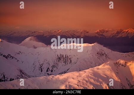 Harte Winde wehen Schnee über die zerklüfteten Bergrücken und Gipfel der South Chilkat Mountains und erleuchten intensive, orange Farben eines Winteruntergangs. Die... Stockfoto