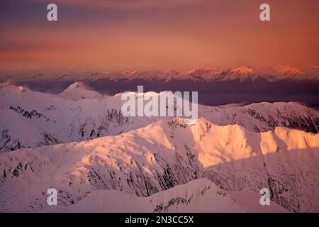 Harte Winde wehen Schnee über die zerklüfteten Bergrücken und Gipfel der South Chilkat Mountains und erleuchten intensive, orange Farben eines Winteruntergangs. Die... Stockfoto