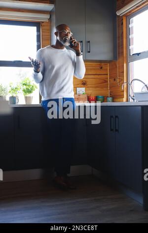 Ein afroamerikanischer, glatzköpfiger Senior-Mann, der sich am Küchentisch in der Blockhütte über Handy unterhält Stockfoto