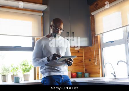 Ein afroamerikanischer, glatzköpfiger Senior-Mann, der ein digitales Tablet benutzt, während er in der Blockhütte am Küchenschalter steht Stockfoto