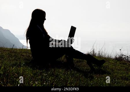 Frau, die Freiberuflerin, die auf einer hohen Klippe über dem Meer sitzt und aus der Ferne arbeitet Stockfoto