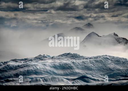 Eisberg im Vordergrund wirkt wie eine Bühne für windgepeitschten Schnee, der an Bergen entlang der Eisberggasse auf dem Weg in die westliche Antarktis ... vorbeiweht Stockfoto