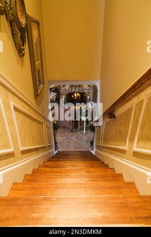 Eine Kiefernholztreppe führt hinunter ins Erdgeschoss mit zwei gepolsterten Sesseln vor dem Kamin aus Naturstein mit Weihnachtsdekoration. Stockfoto