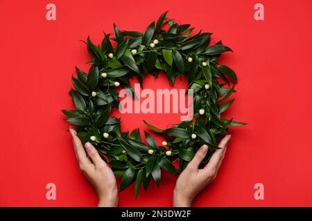 Frau mit wunderschönem Mistelkranz auf rotem Hintergrund, Draufsicht. Traditionelle Weihnachtsdekor Stockfoto