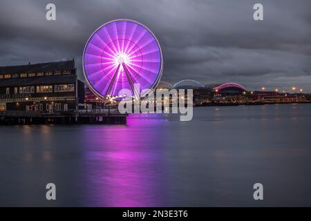 Ein bewölkter Abend am Ufer von Seattle mit Blick auf das Seattle Great Wheel, das sich von der Elliott Bay reflektiert, mit Lumen Field und T-Mobile Park beleuchtet ... Stockfoto