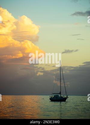 Ein einsames Segelboot, das vor der Küste vor Anker liegt und das goldene Licht des Sonnenuntergangs reflektiert auf dem türkisfarbenen Wasser des Südpazifiks; Tahaa, Französisch-Polynesien Stockfoto