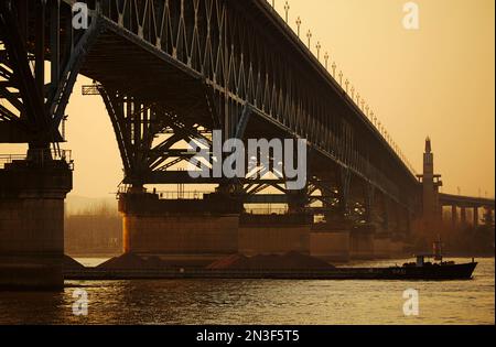 Silhouette des Schiffs, das unter der Yangtze-Brücke durch den Yangtze-Fluss in der Abenddämmerung fährt; Nanjing, Provinz Jiangsu, China Stockfoto