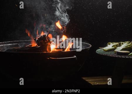 Brennholz brennt in einem runden Barbecue in der Nacht. Dunkler Hintergrund. Vorbereitung zum Kochen von Speisen auf dem Grill. Stockfoto