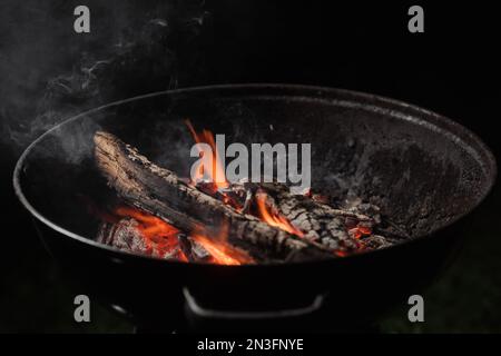 Brennholz brennt in einem runden Barbecue in der Nacht. Dunkler Hintergrund. Vorbereitung zum Kochen von Speisen auf dem Grill. Stockfoto