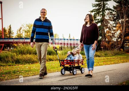 Ein Vater und eine Mutter, die während der Herbstsaison ihre kleinen Kinder in einem Wagen in einem Stadtpark ziehen, und ihr kleines Mädchen hat Down-Syndrom Stockfoto