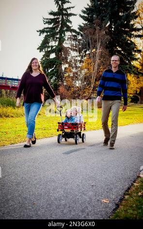 Ein Vater und eine Mutter, die während der Herbstsaison ihre kleinen Kinder in einem Wagen in einem Stadtpark ziehen, und ihr kleines Mädchen hat Down-Syndrom Stockfoto