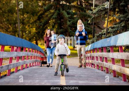 Junge Familien, die in der Herbstsaison eine Brücke über einen Fluss in einem Stadtpark überqueren, und ihre kleine Tochter hat Down-Syndrom Stockfoto