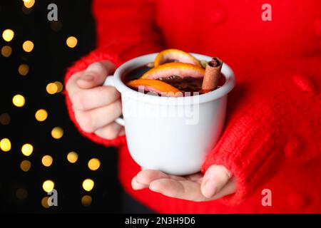 Eine Frau, die eine Tasse Glühwein gegen verschwommenes Licht hält, Nahaufnahme Stockfoto