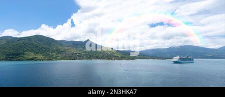 Panoramafoto von Ilha Bela an einem sonnigen Tag mit einem Schiff vor Anker. Im Hintergrund ein Regenbogen. Stockfoto