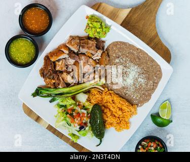 Ein Blick von oben auf Carnitas Teller auf einem Holzbrett mit gekühlten Bohnen, Reis, Salsa und Limette Stockfoto