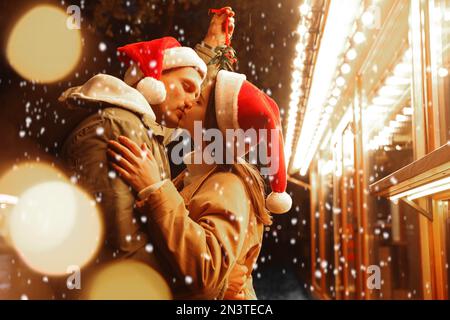 Glückliches Paar küsst sich unter Mistelzweigen draußen an verschneiten Abenden, Bokeh-Effekt Stockfoto