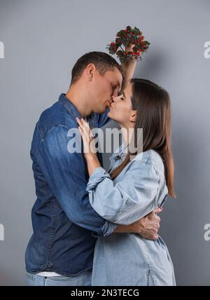 Ein glückliches Paar küsst sich unter dem Mistelkranz auf grauem Hintergrund Stockfoto