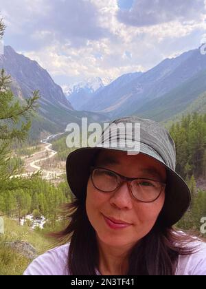 Ein asiatisches Mädchen mit Brille und Hut macht ein Selfie vor dem Hintergrund eines Panoramas aus Bergen und einem Fluss mit Gletschern und Schnee in Altai Stockfoto